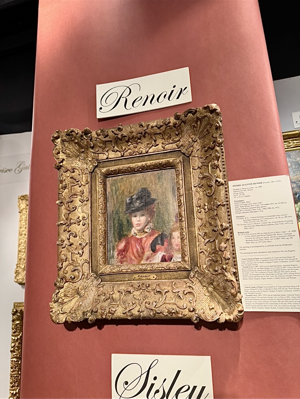 Ornate Gold frame surrounds small Renoir painting of a hatted woman in pinkish red.