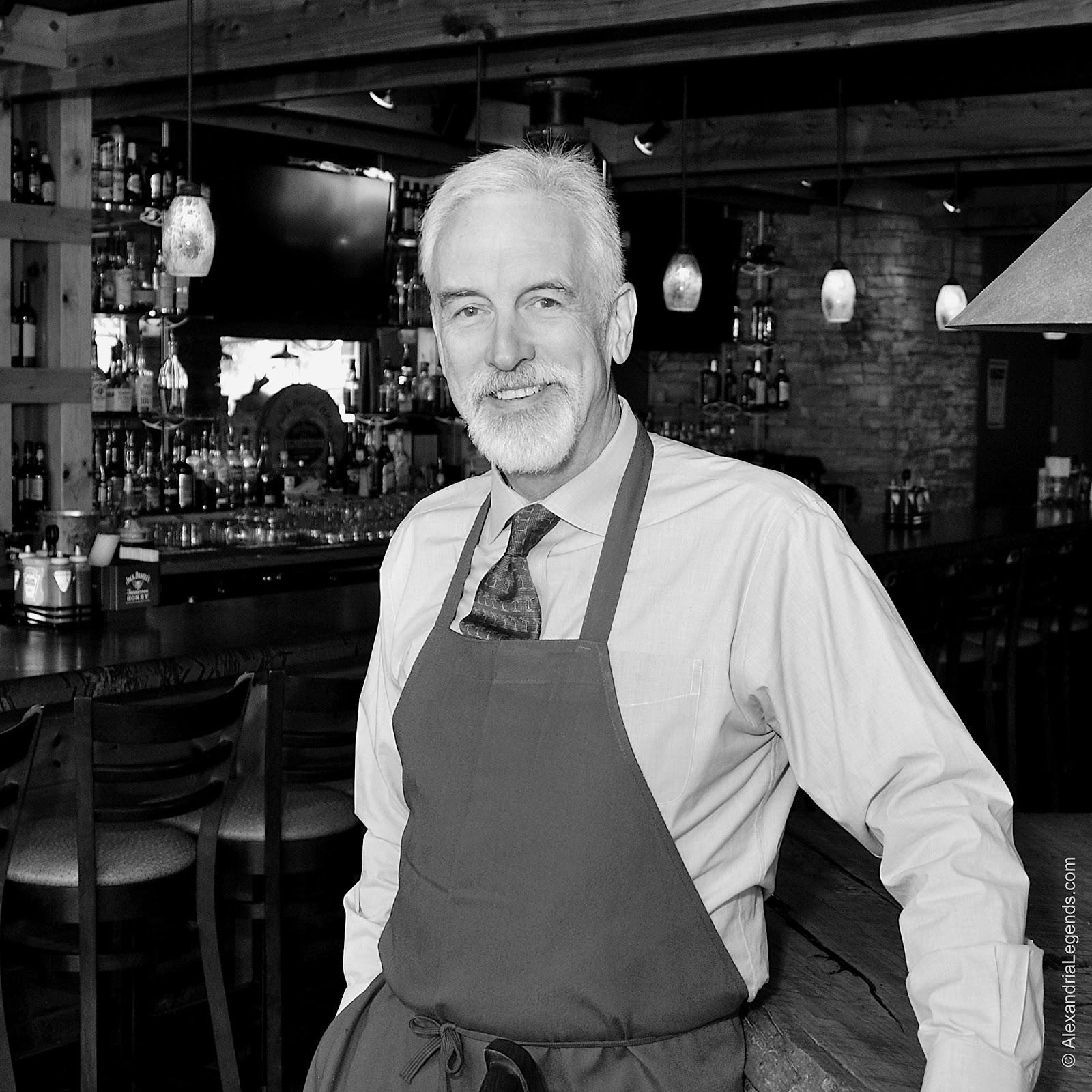 Man with white hair and beard wearing shirt and tie with chef's apron.