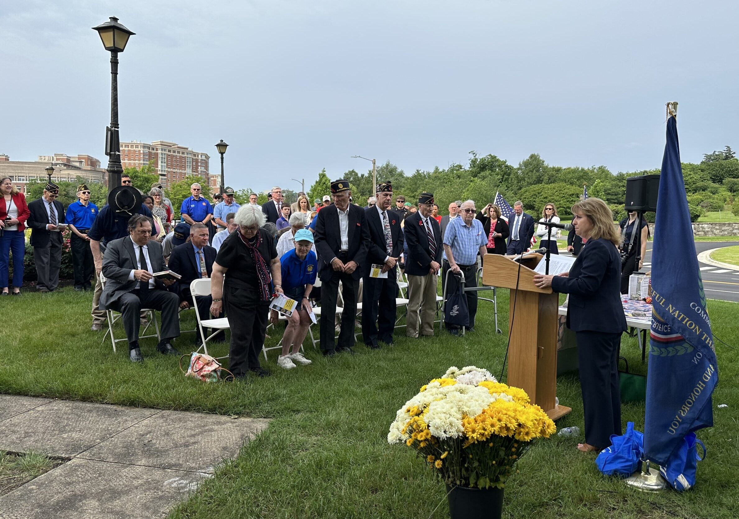 A crowd gathers on a green lawn and listens to aa woman at a podium.