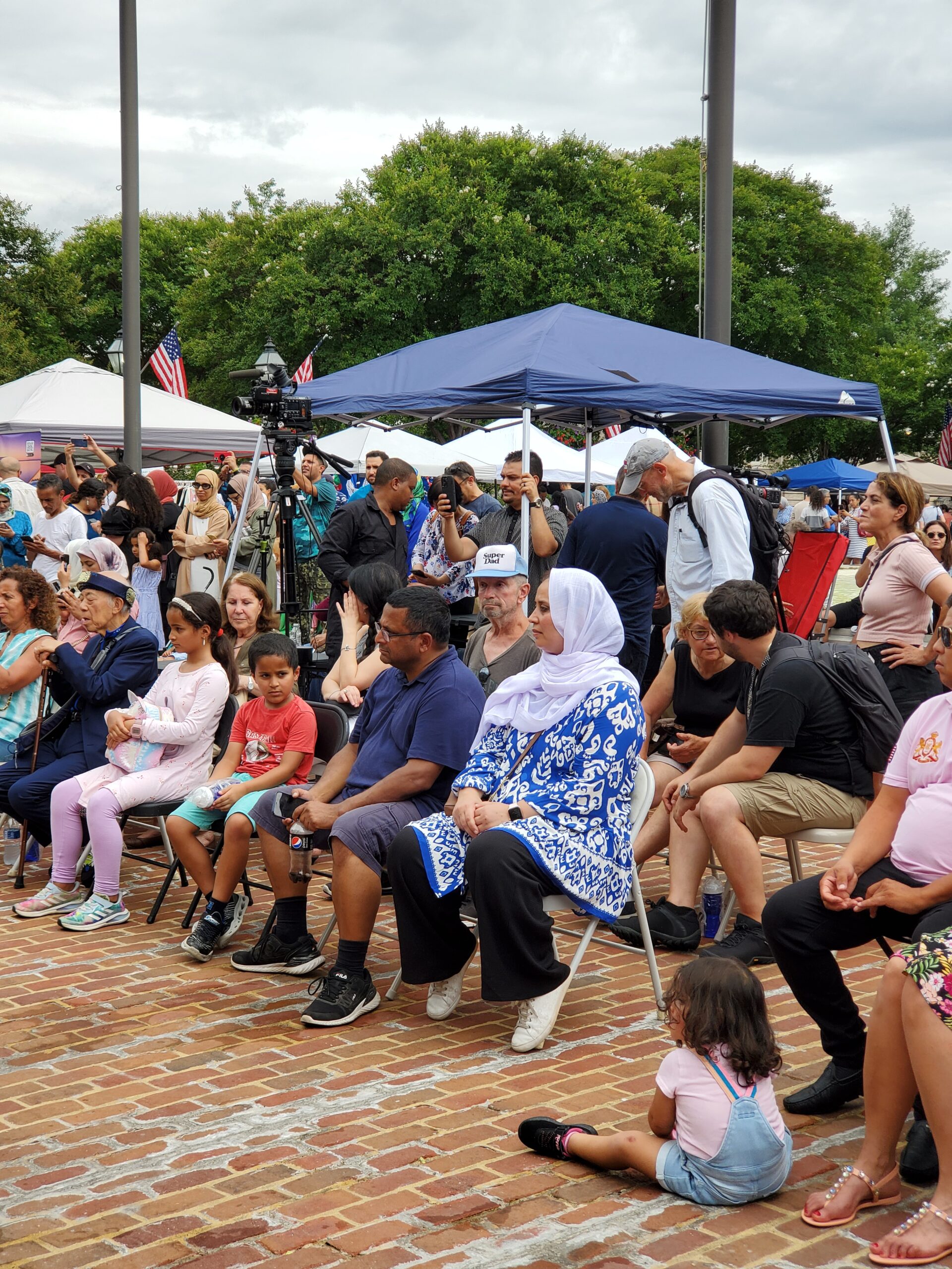 People sitting and standing in a crowd with vendor tents in background.