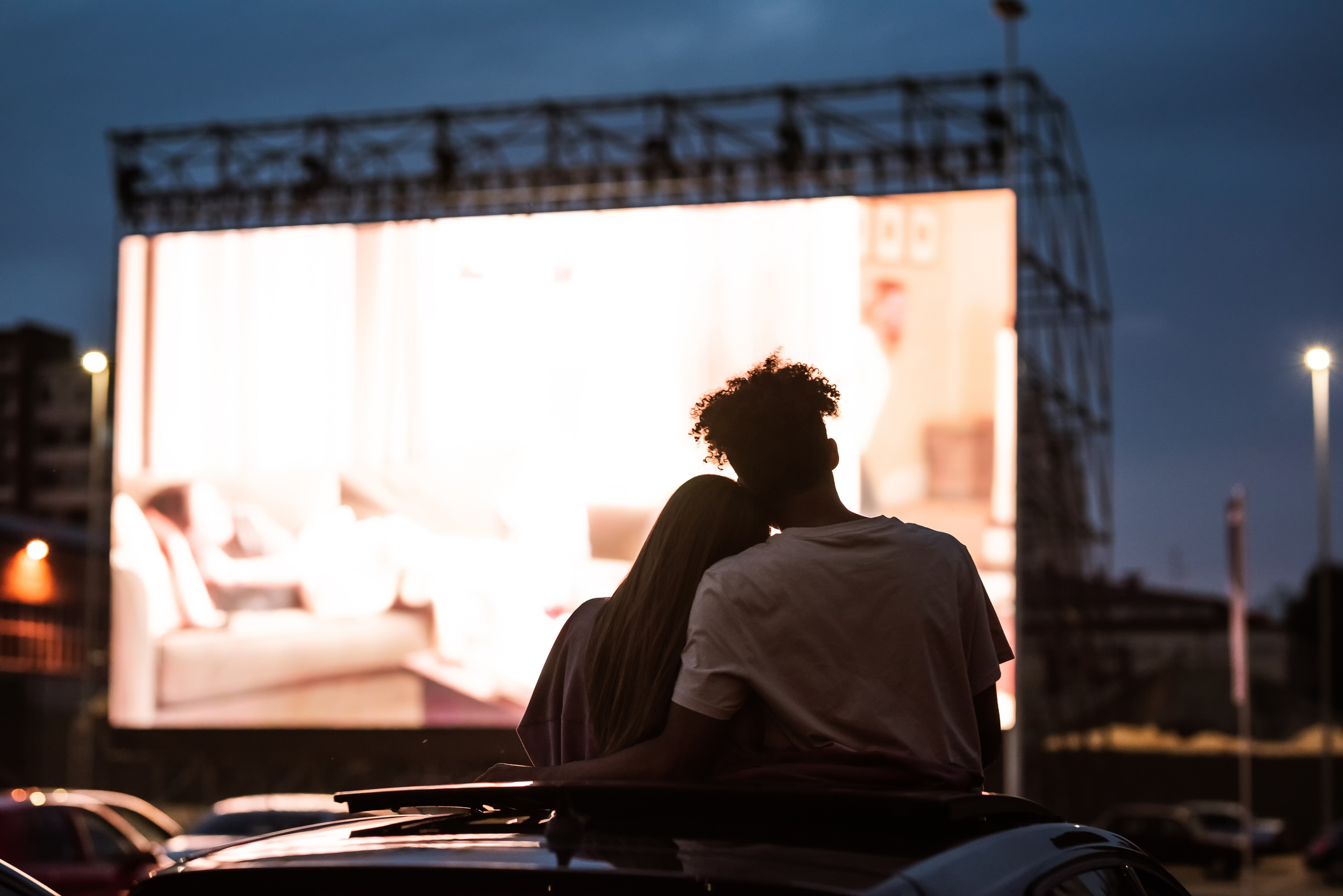 Silhouetted view of attractive young couple, boy and girl embracing, spending time together, sitting in the car while watching a movie in a drive in cinema Silhouetted view of attractive young couple, boy and girl embracing, spending time together, sitting in the car while watching a movie in a drive in cinema
