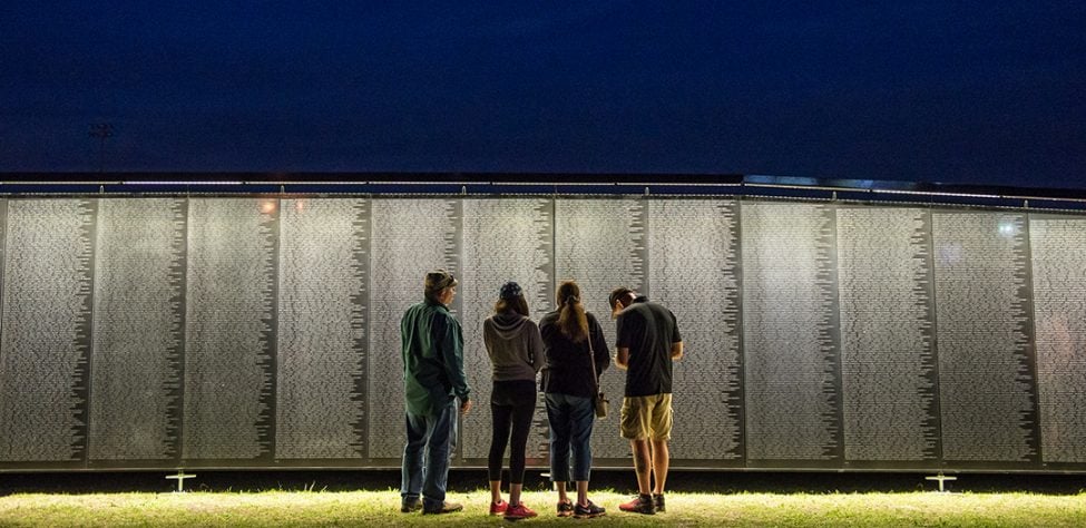 Four people looking at The Wall that Heals, the three-quarter sized replica of the Vietnam Veterans Memorial. (Photo: Vietnam Veterans Memorial Fund) Four people looking at the Vietnam Veterans Memorial Replica at night with the light glowing on the wall