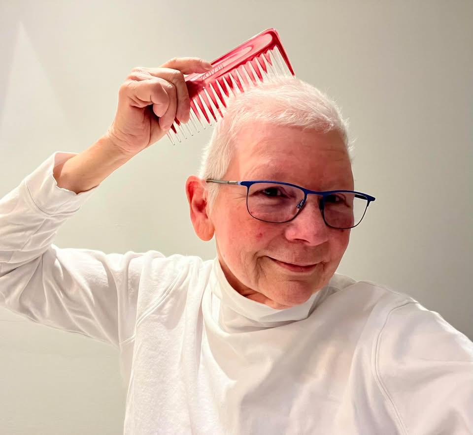 Lee Moody smiles while holding a bright red comb above her short white hair during cancer treatment, wearing blue glasses and a white top against a simple light background. Lee Moody smiles while holding a bright red comb above her short white hair during cancer treatment, wearing blue glasses and a white top against a simple light background.
