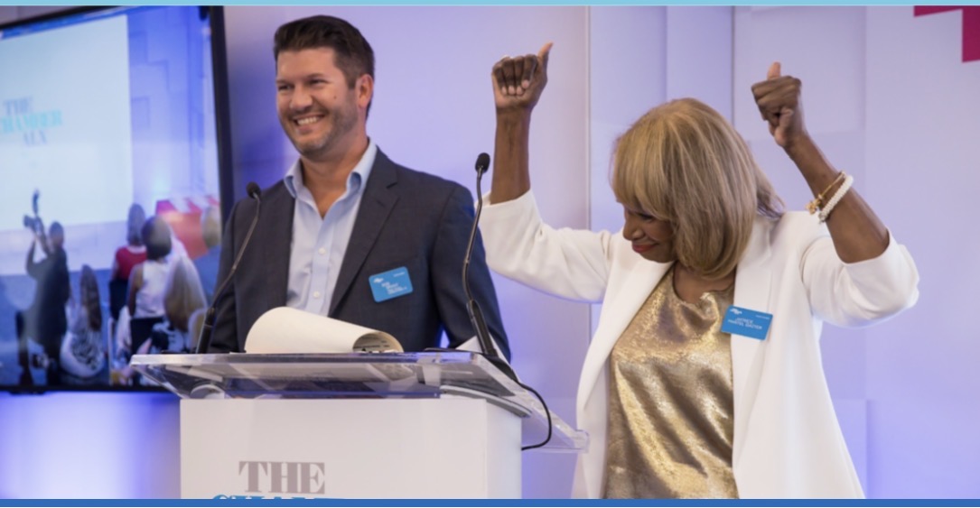 Two speakers stand at a podium during a Chamber ALX event, smiling and celebrating as one woman raises both arms enthusiastically in front of an audience.