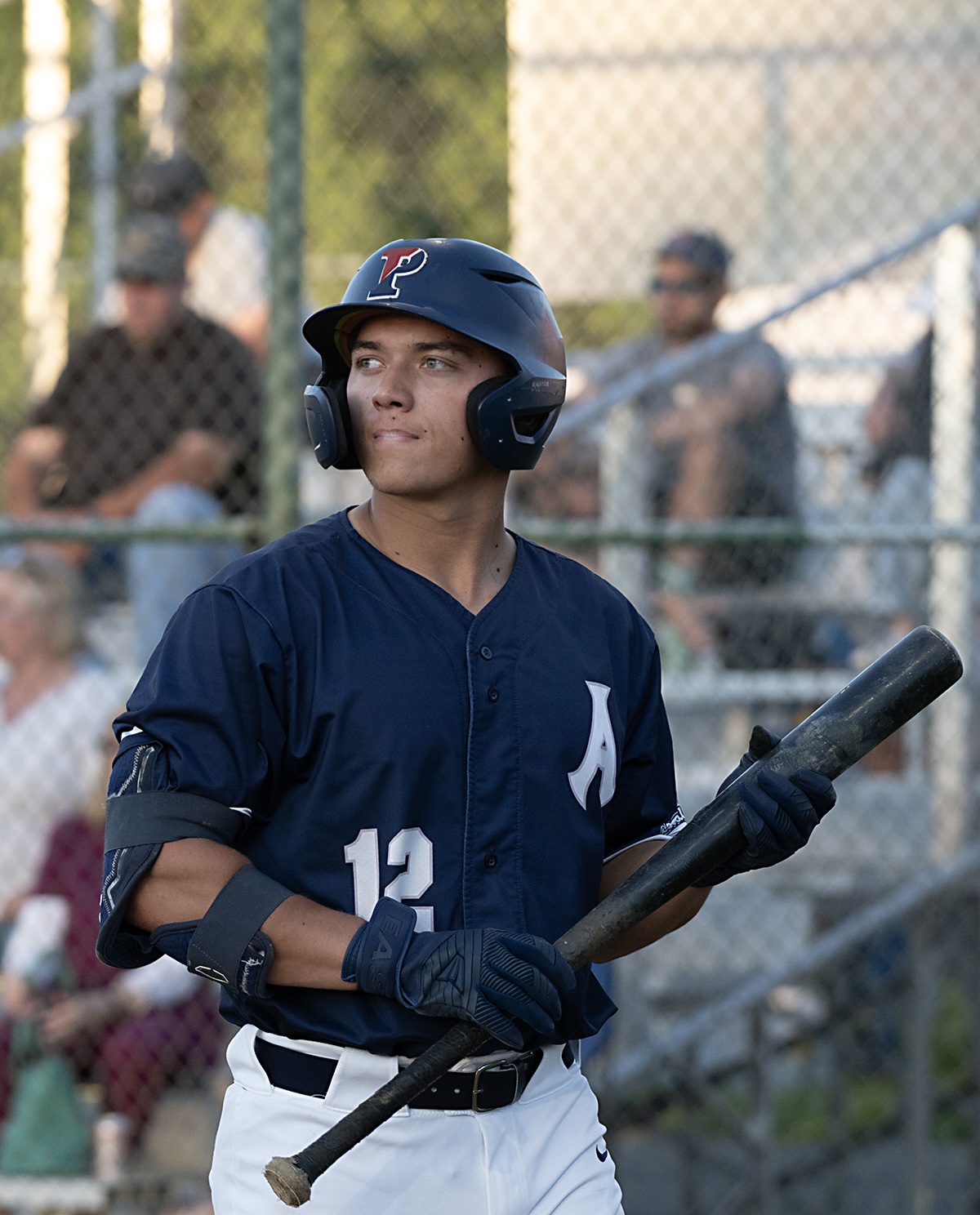 Jack Warner (University of Pennsylvania) waits on deck during an Alexandria Aces game at Frank Mann Field. Photo by Mark Briscoe/The Zebra Press. Alexandria Aces batter Jack Warner, wearing jersey number 12, waits on deck with a bat in hand during a summer collegiate baseball game. Fans watch from the bleachers behind him.