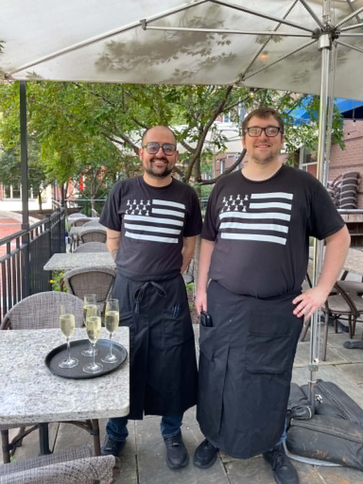 Andres Henao and Johnny Houllahan stand on the Maison Breizh patio beside a tray of champagne during the July 15 ribbon-cutting celebration.