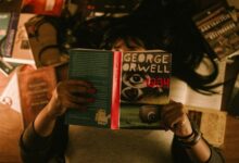 A person holds a copy of George Orwell's 1984 while reading indoors, surrounded by books.