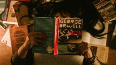 A person holds a copy of George Orwell's 1984 while reading indoors, surrounded by books.