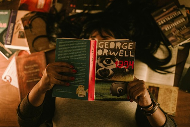 A person holds a copy of George Orwell's 1984 while reading indoors, surrounded by books.
