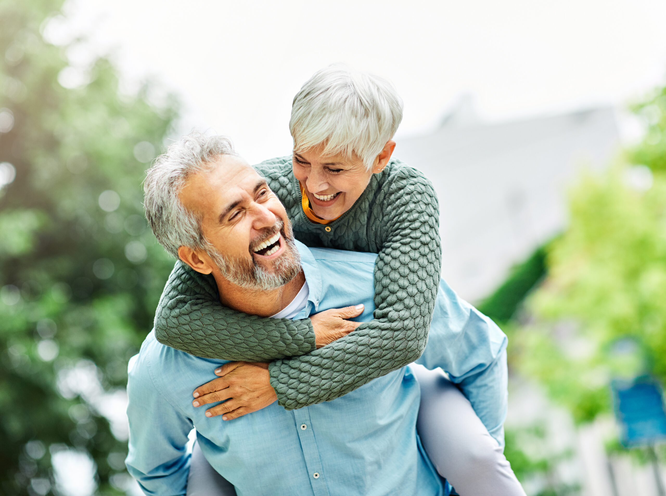 A smiling older couple shares a playful piggyback ride outdoors in a park, laughing together on a sunny day.