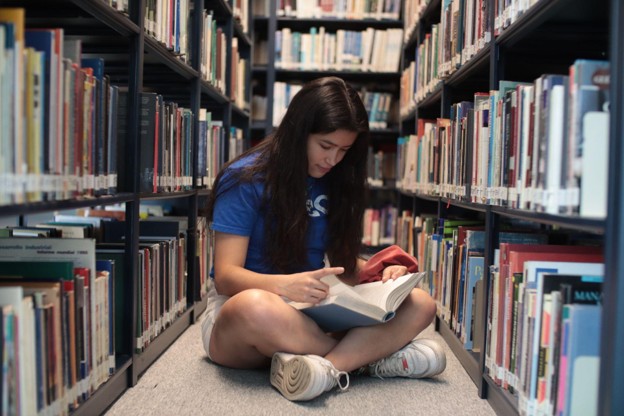 Student Reading in Library Aisle A young student sits on the floor between library bookshelves reading a book.