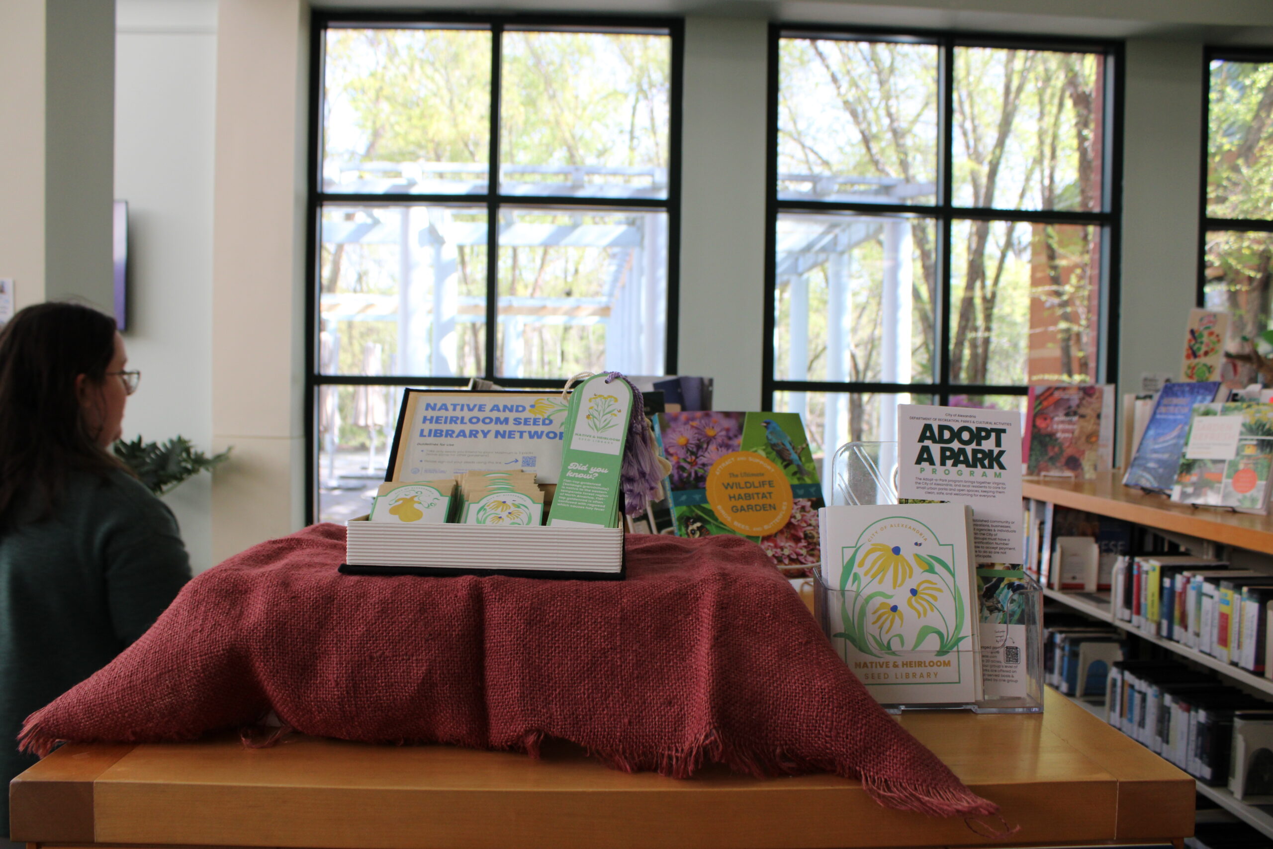 IMG 0304 Natural Resources Manager Katrina Napora next to the Native and Heirloom Seed Library Display in Charles E. Beatley Regional Library. Surrounding educational resources about gardening are available to check out, and the native reading garden is visible from the windows. Photo by Leah Sly
