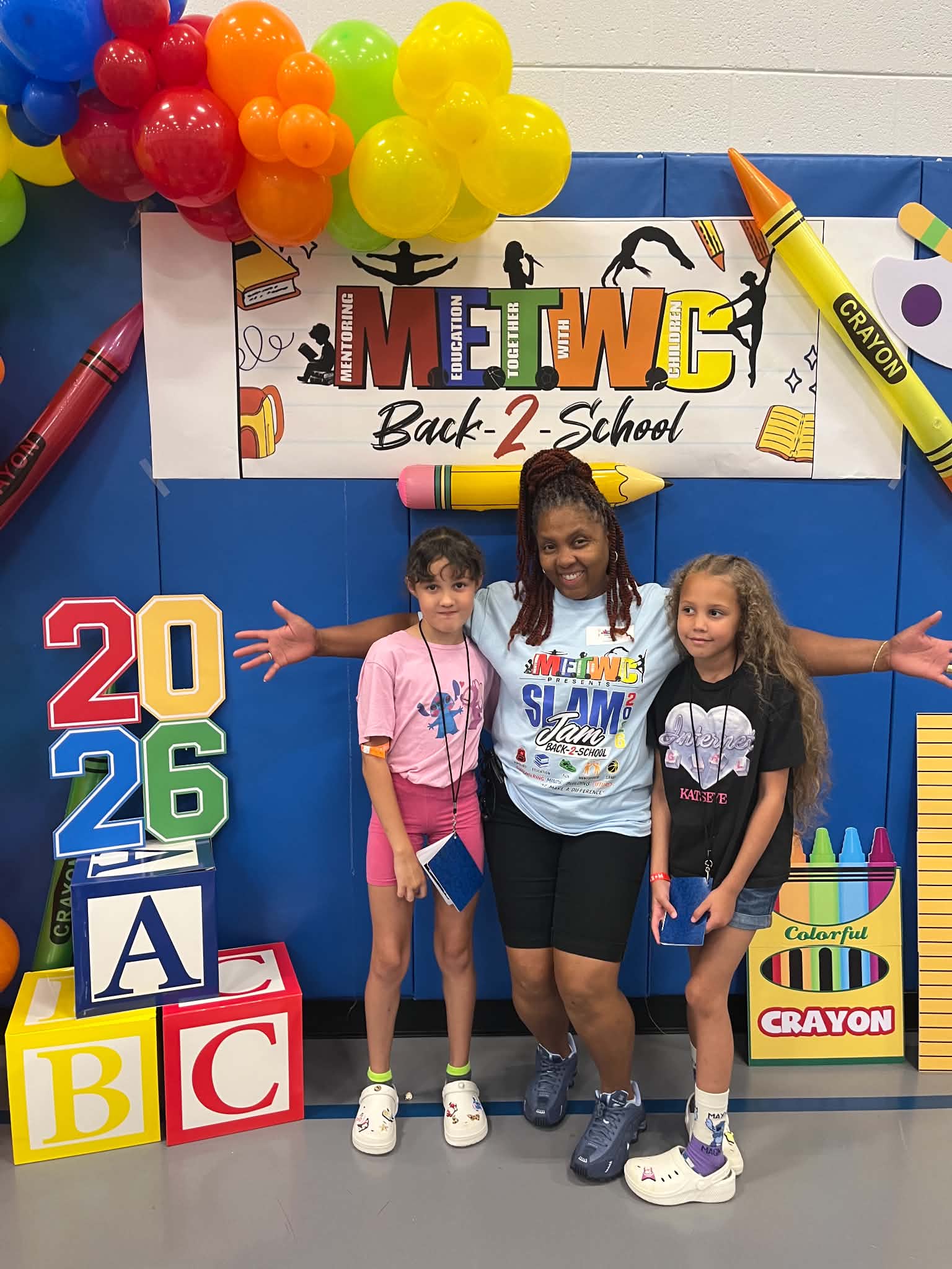 METWC Director Felecia Parks stands between Kaia Davis, 8, and Lani Davis, 7, in front of a colorful Back 2 School Slam Jam display at Patrick Henry Recreation Center.