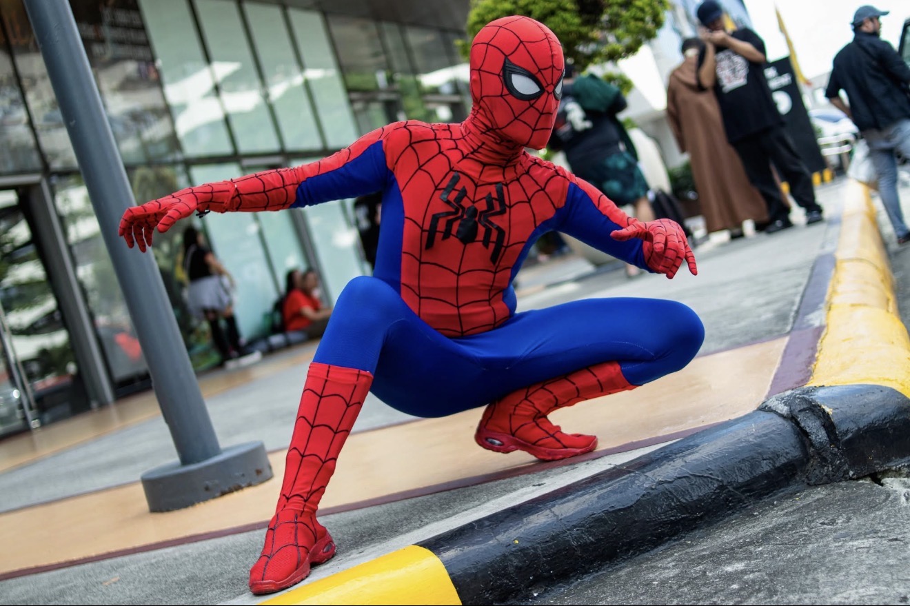 Cosplayer Cyan Blur crouches in a red-and-blue Spider-Man costume beside a curb.