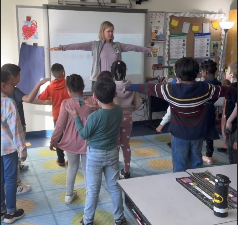 Teacher and elementary school students stretch their arms during a classroom movement activity.