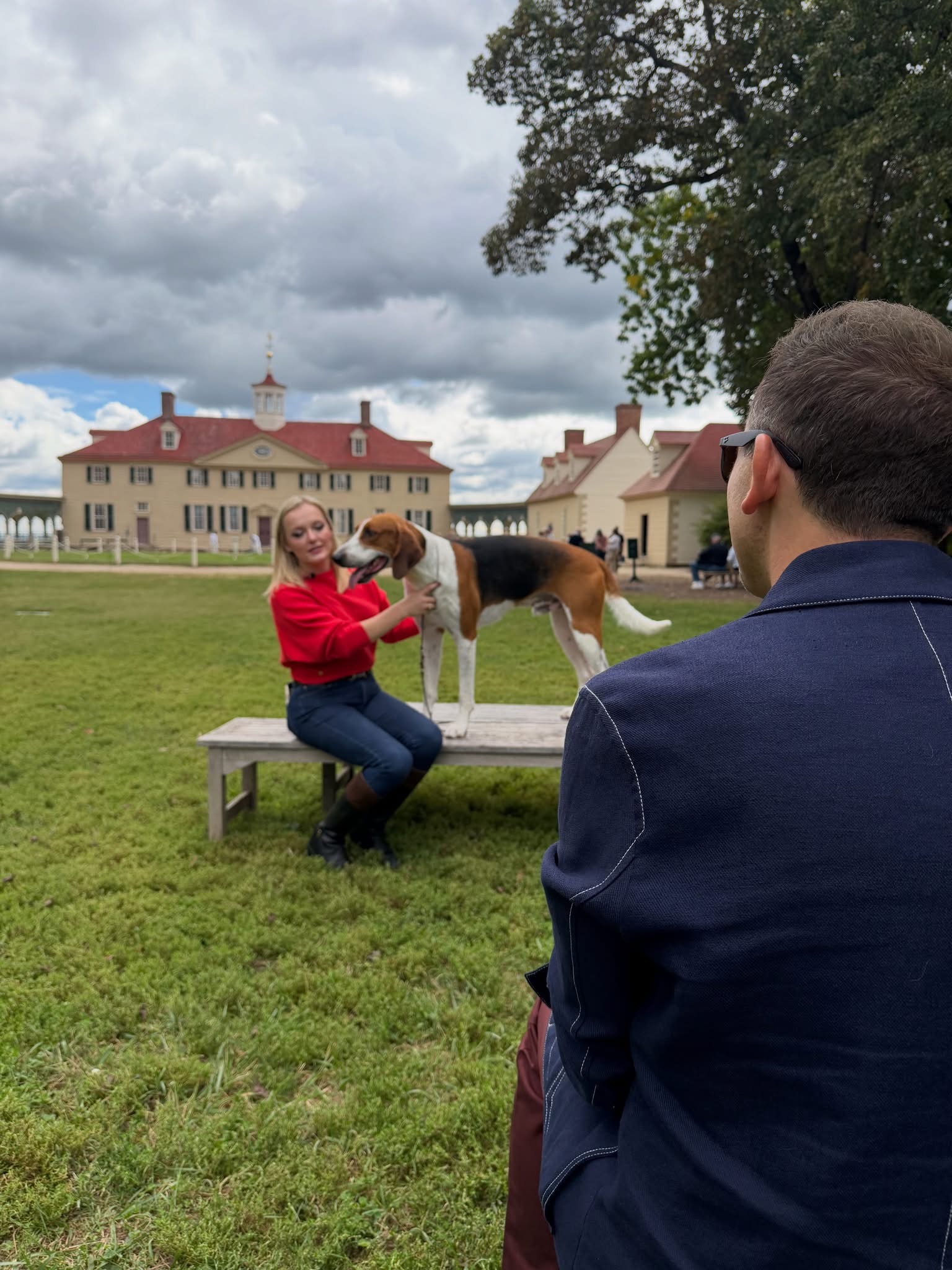 A woman in a red shirt poses George the American foxhound on a wooden bench at Mount Vernon while a man stands in the foreground during an interview setup.