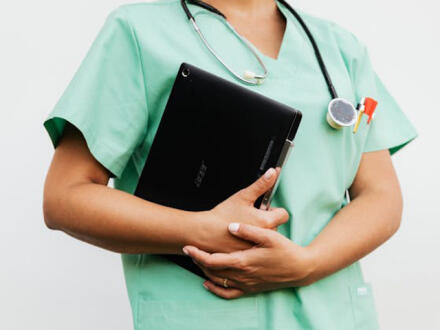 Person in mint-green medical scrubs wearing a stethoscope and holding a black laptop.