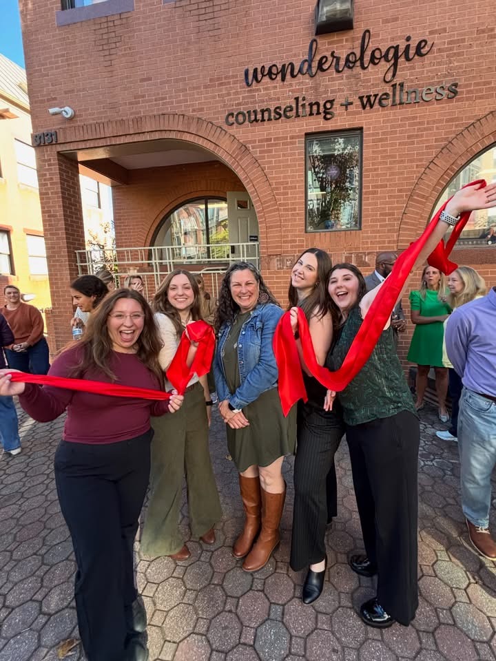 Five smiling attendees celebrate with a red ribbon outside Wonderologie’s brick building in Del Ray.