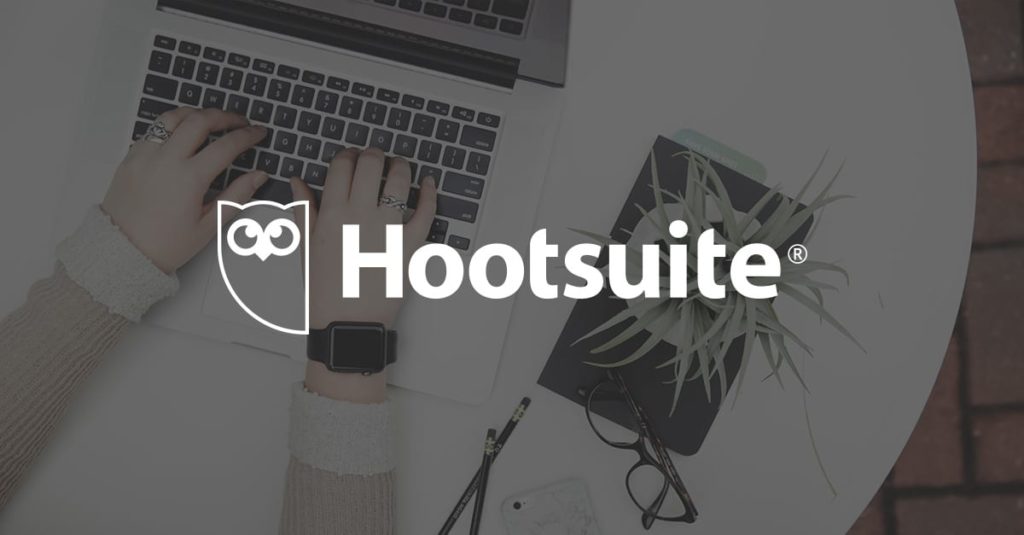 The Hootsuite logo over a background of a person typing on a laptop with a plant, eyeglasses, and a notebook on a white table.
