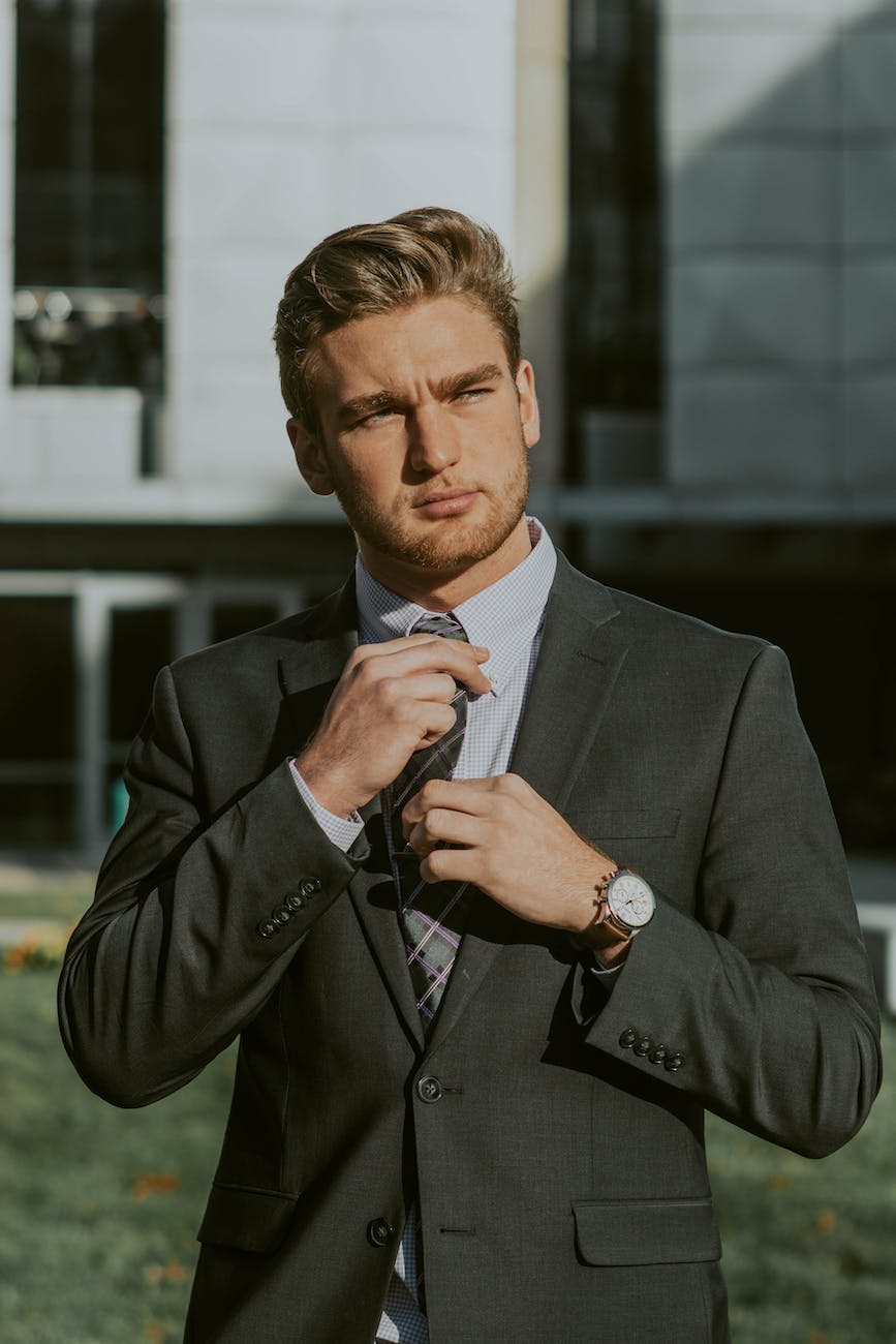 A man in a dark suit stands outdoors, adjusting his tie with one hand. He is looking into the distance with a serious expression. A building with large windows is in the background.