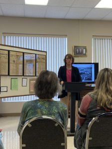 Debra Herndon, museum director, speaks at podium, with newspaper clippings on a display to her left