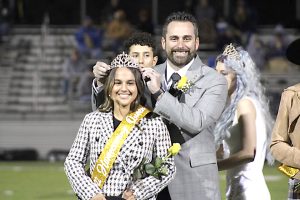 Keri in checker-print blazer and yellow sash, being crowned on the field, both she and the principal beaming