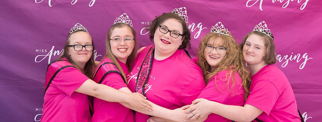 girls in pink shirts, sashes and tiaras hug and smile for the camera