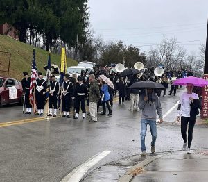 People line up at parade with flags and instruments; two onlookers have umbrellas.