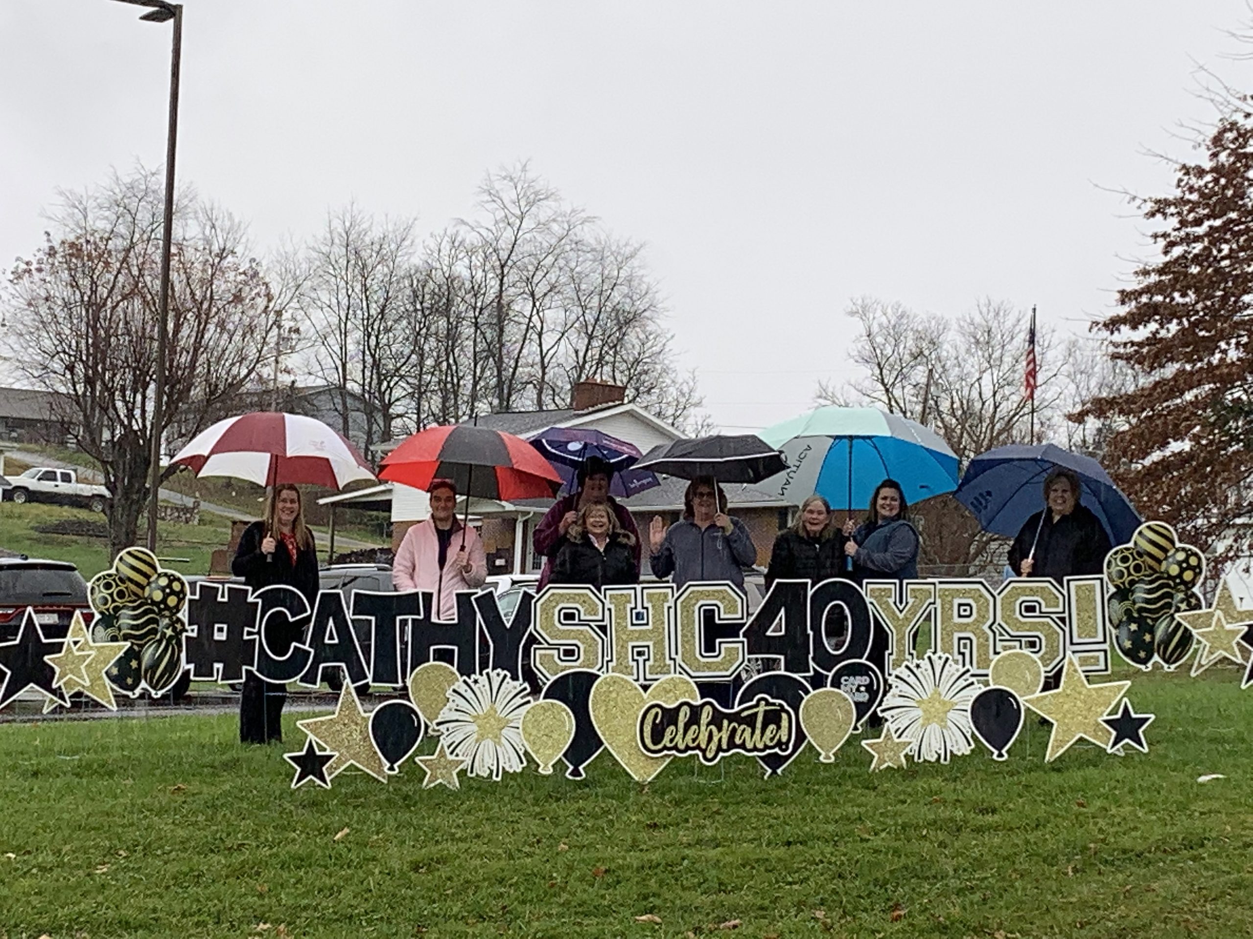 Group of Shinnston Healthcare workers, holding umbrellas, stand around sign that says CATHYSHC40YRS! Underneath it says Celebrate! 