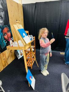 Piper in front of an easel holding up a paintbrush 