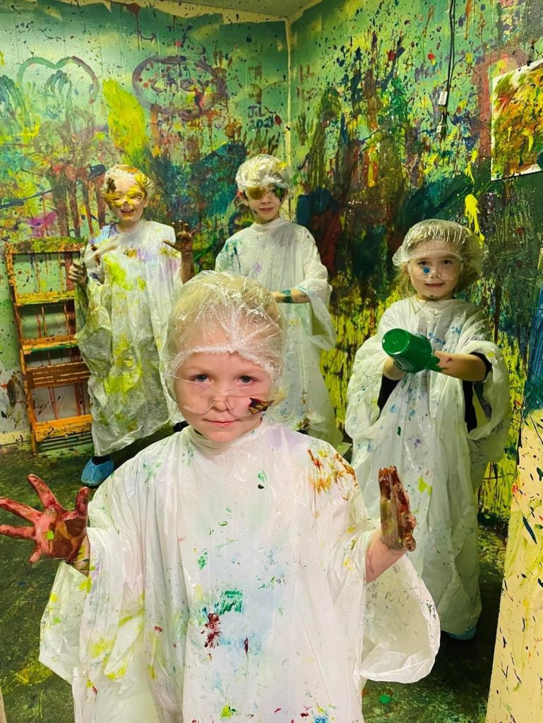 kids are splattered in paint, surrounding by walls splatter in paint, Piper is in front looking very serious about the task