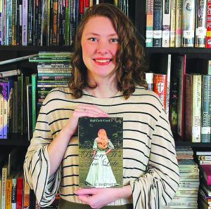 She is holding her book in front of a bookcase. She is smiling.