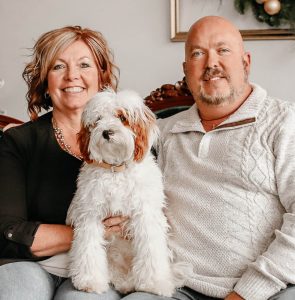 They are sitting on the couch smiling with a fluffy white dog in between.