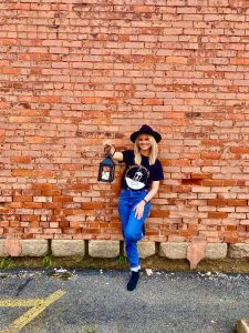 She is standing against a brick wall. She is smiling and holding out a lantern.