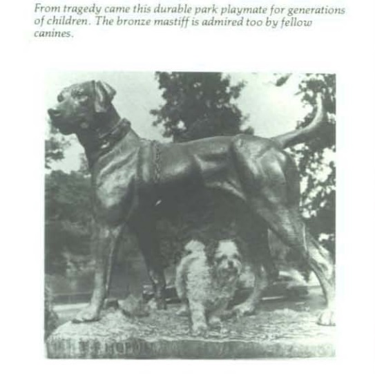 Photograph of a bronze mastiff statue with a toy breed dog standing under it.