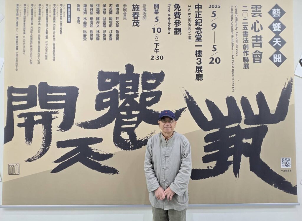 ▲雲心書會指導老師施春茂教授在所書寫的展覽主題「藝饗天開」前留影,書體可謂大器軒昂,令人驚艷。(記者 辛澎祥/攝)