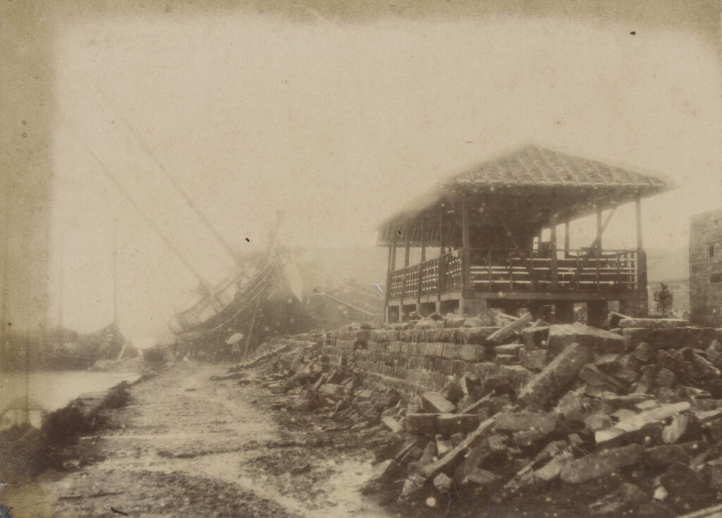 1898「戊戌大水災」照片標示大稻埕淡水河岸，被大水沖垮的石造堤防，堤防上的木屋因位置較高而未受損，後方則有一艘大船被洪水沖上堤岸而擱淺倒於一旁。（圖/國家攝影文化中心提供）
