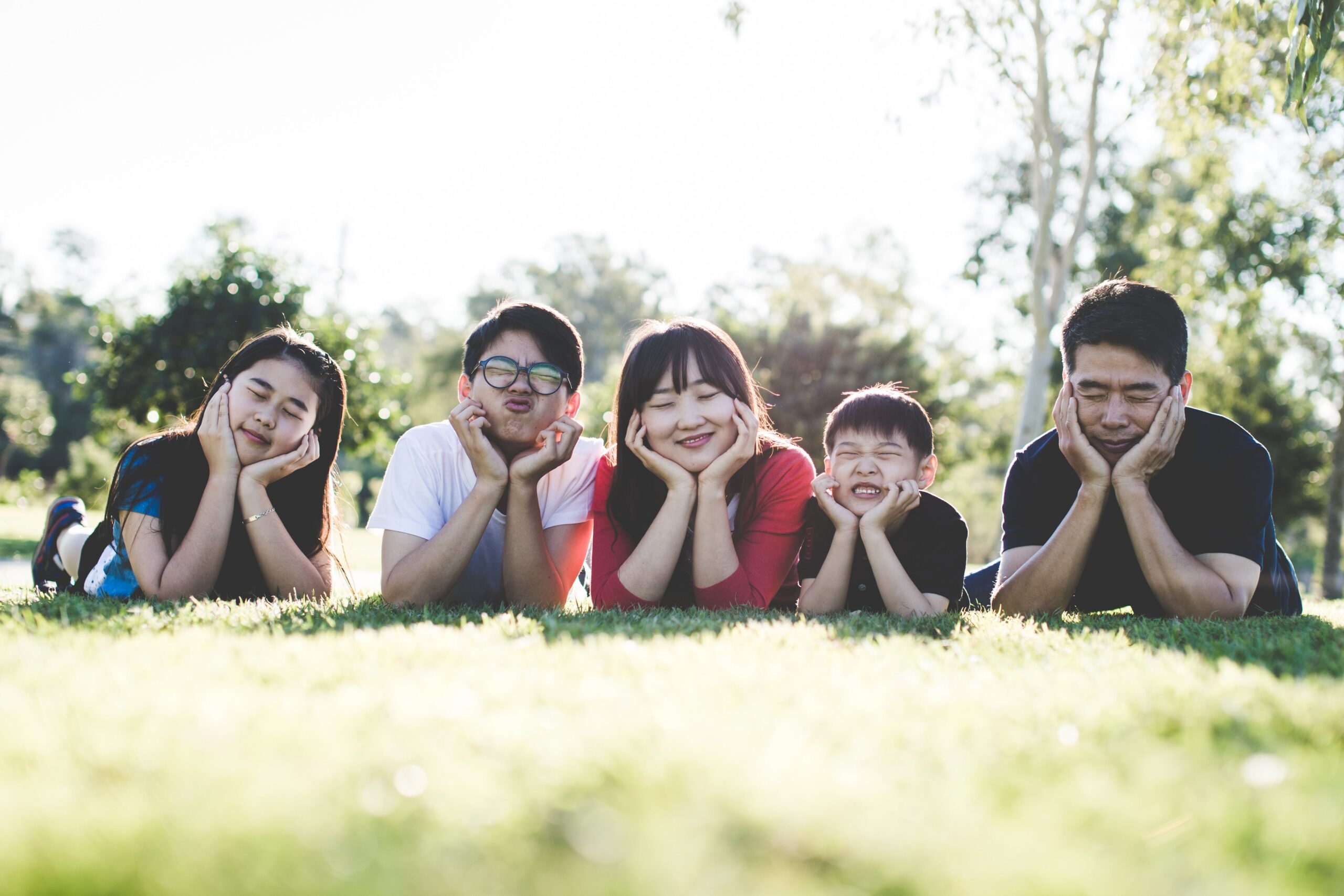 an asian family of 2 adults and three children lying on the grass with their hands under their chins holding their heads up