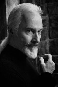 Rick Baker, now 68 years old. He stares into the camera and has his iconic long, white hair pulled into a pony tail.
