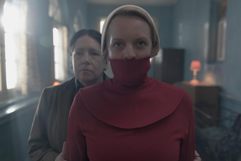 June wearing a mouth veil, which is common to the handmaids in Washington, is being led down a hallway by Aunt Lydia. 