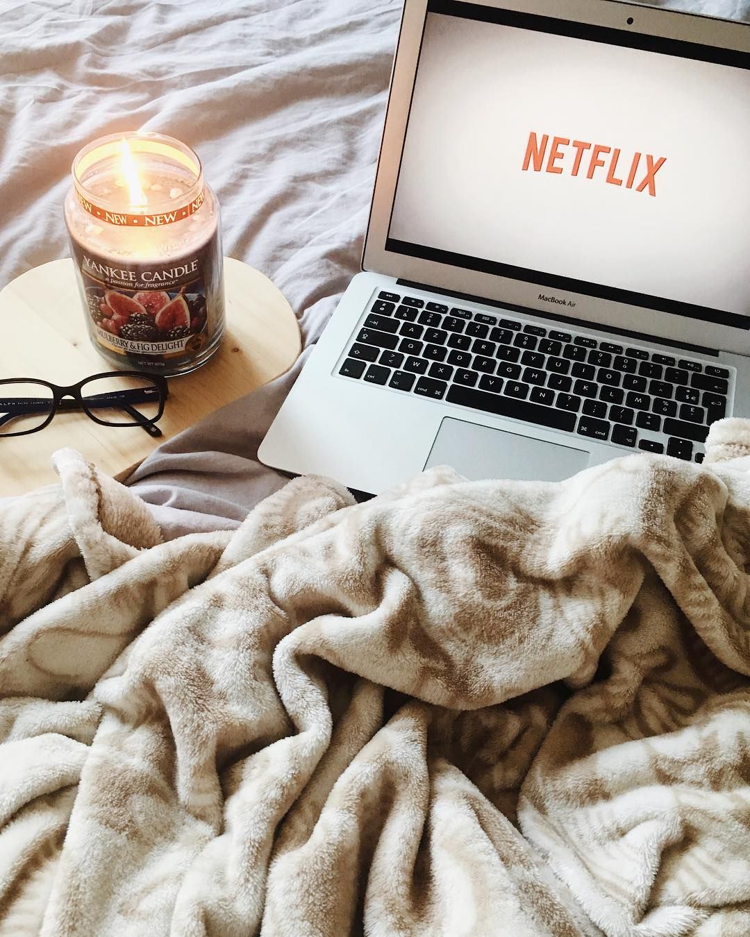 A laptop, candle, and pair of reading glasses sit on top of a rumpled blanket.