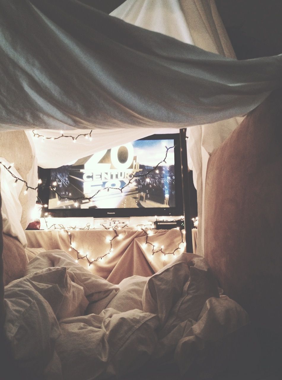 A cozy pillow and blanket fort with string lights and a tv in the background.