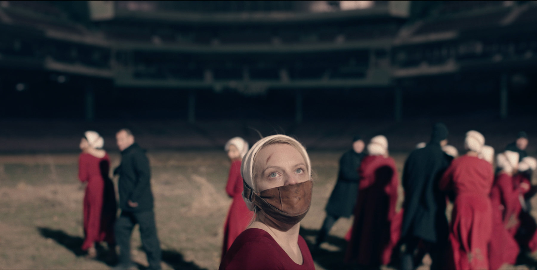 June and other handmaids are being led by guards around an abandoned stadium field. 