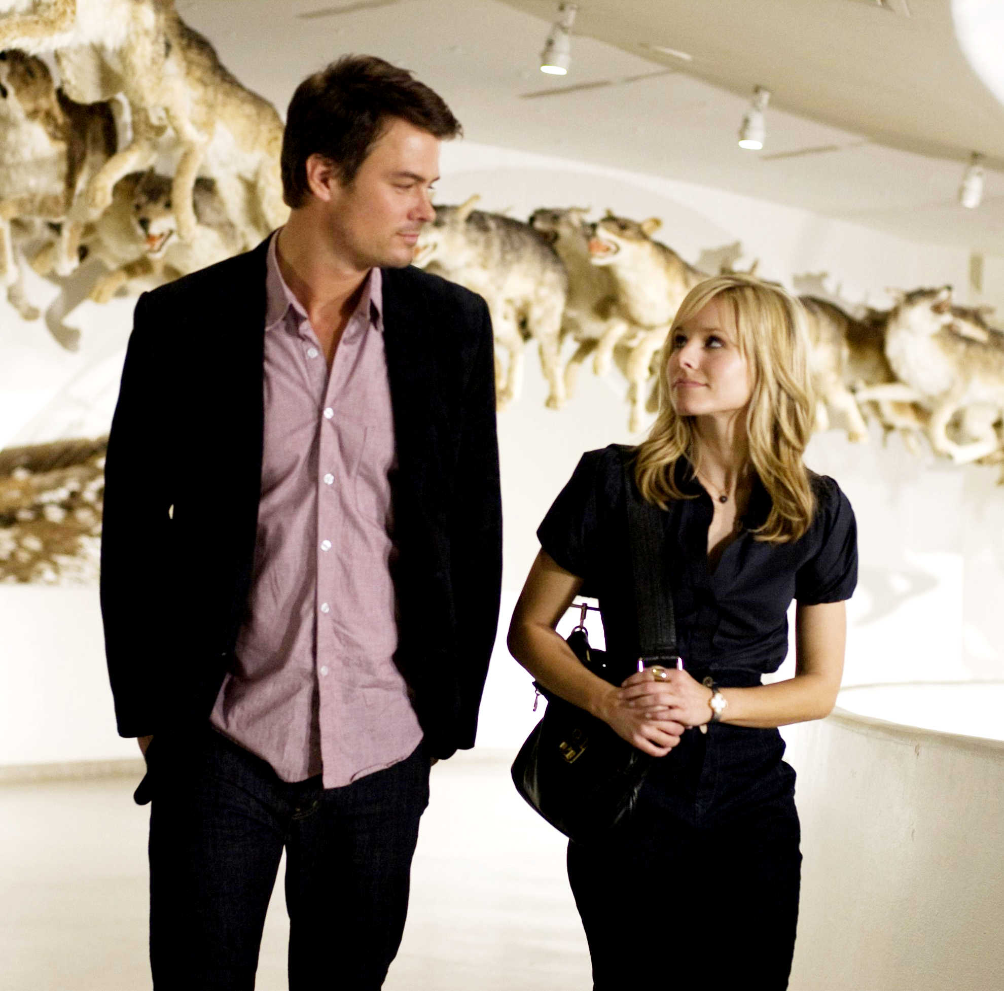 The love interests walk side-by-side in an art gallery while gazing at one another.