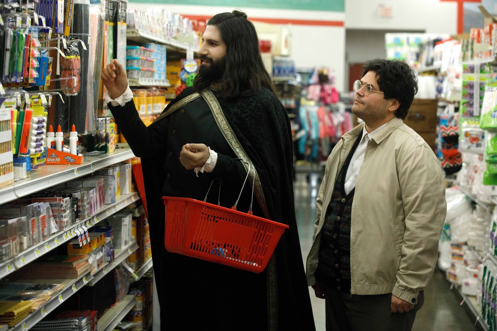 Nandor in full vampire garb and Guillermo in a supermarket. Nandor is holding a shopping basket and looking at craft supplies.