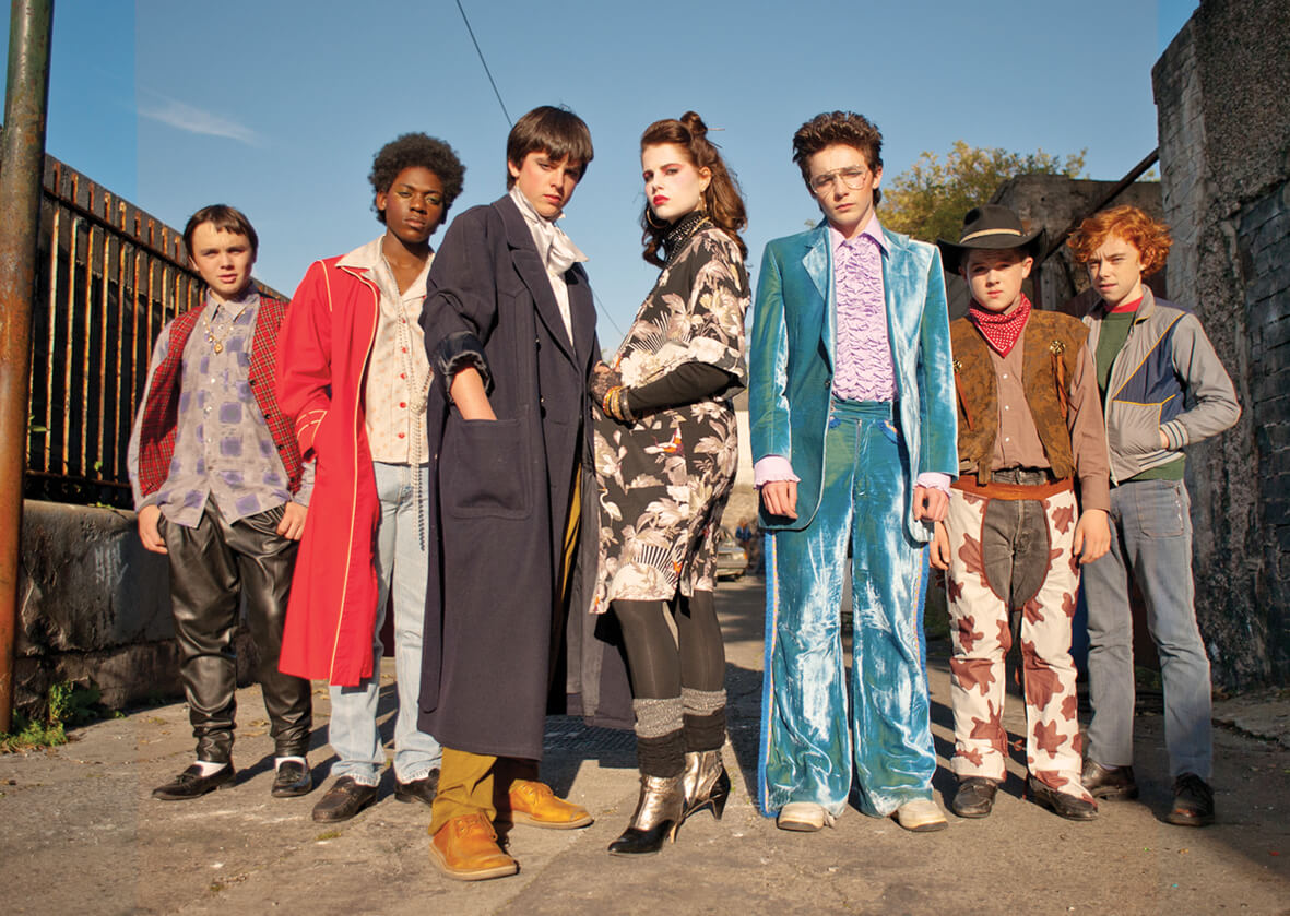 The cast of Sing Street pose in comically uncoordinated outfits during their first music video. Specifically, one of the seven people in the photo wears a velvet 1970's style prom tuxedo, while the person next to him wears a child's cowboy costume.