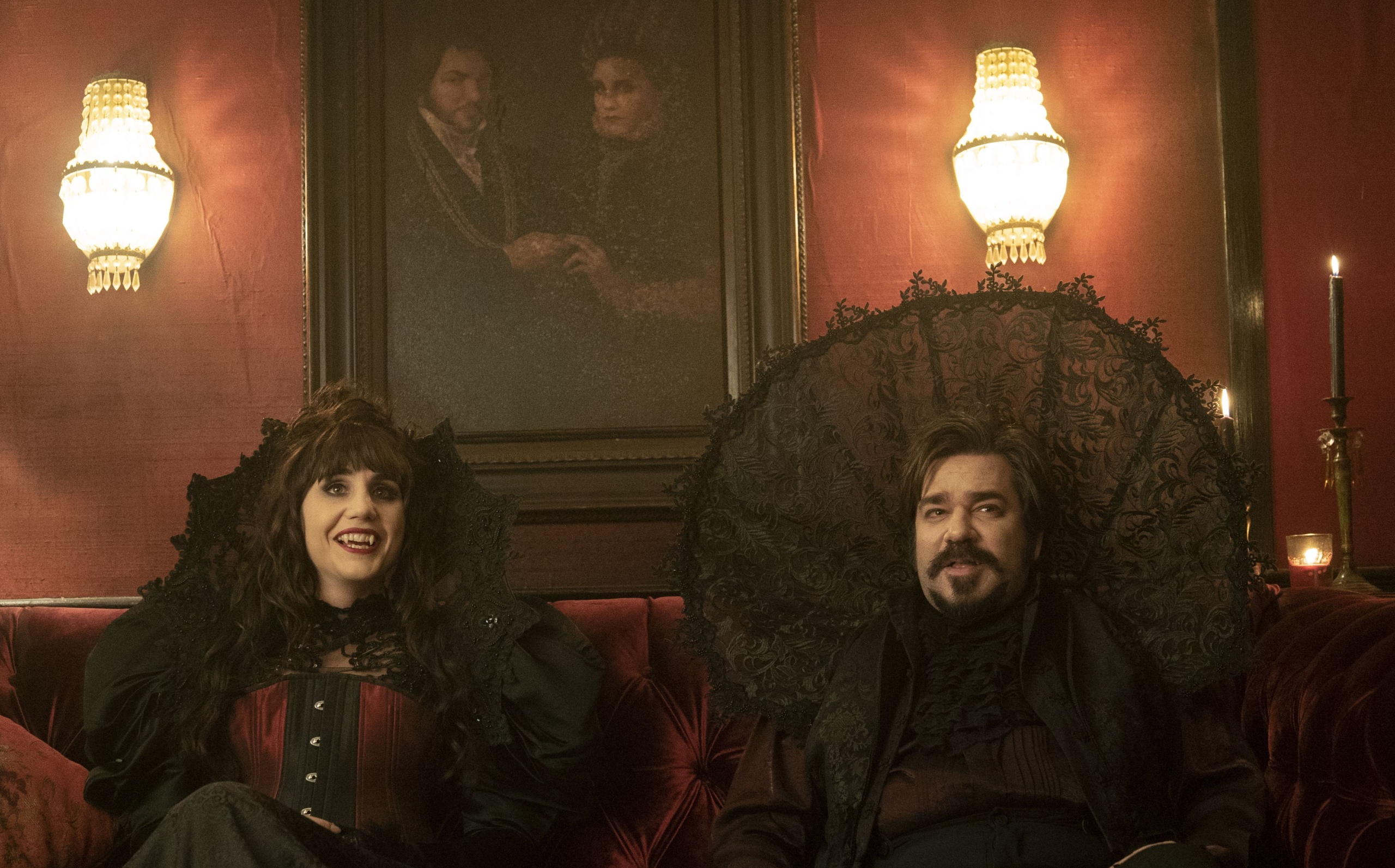 Nadja and Laszlo dressed in gothic outfits with high collars. They sit on a sofa under a portrait of themselves. 