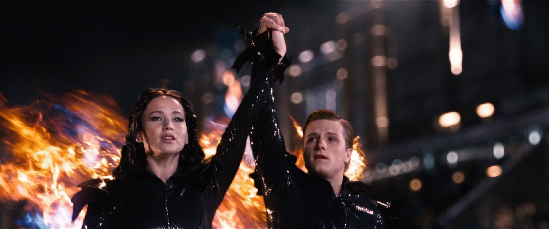 Katniss and Peeta hold hands while fire shoots out from their clothing behind them.