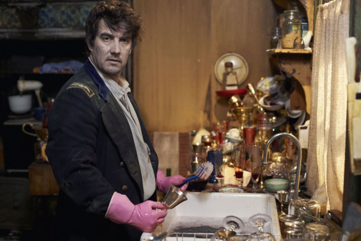 Deacon from What We Do In The Shadows 2014 wearing rubber gloves and washing bloody dishes in a sink.