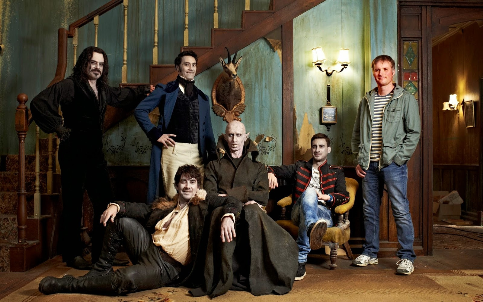 The main cast of the movie What We Do In The Shadows all posing in front of an antique staircase and decor.